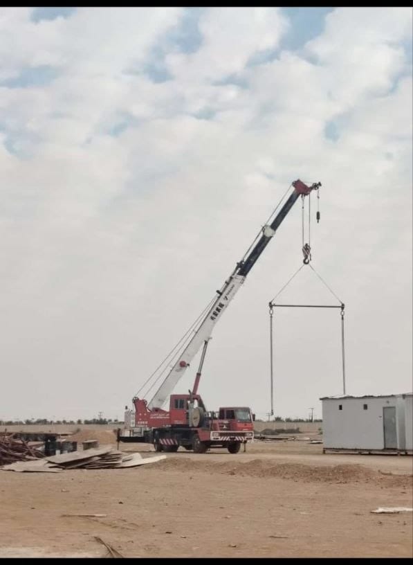 كرين متحرك أحمر يرفع مقطعًا حديديًا طويلاً بحبال وشناكل في موقع مفتوح